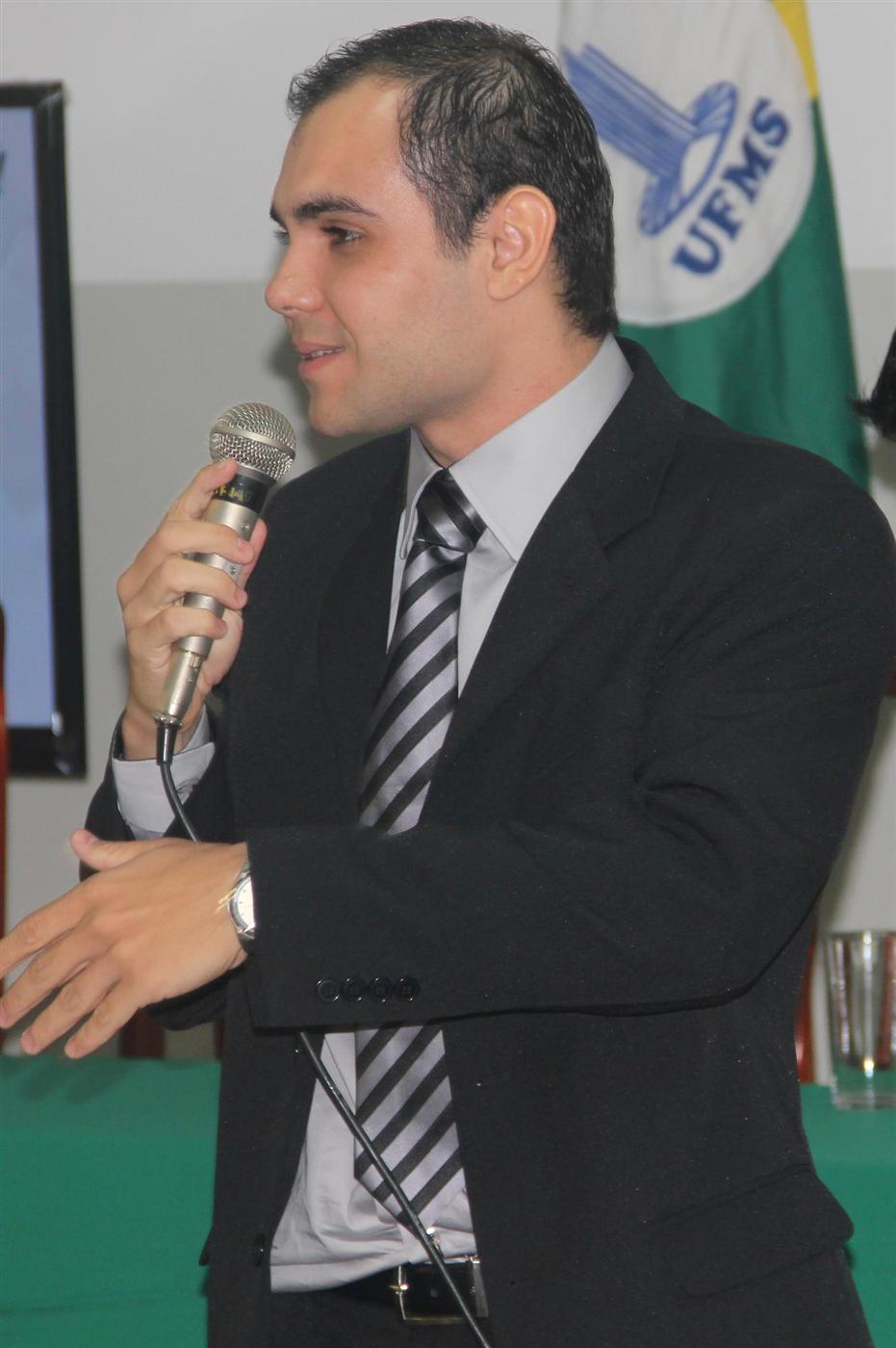 Rodney de terno fala ao microfone em evento, com a bandeira da UFMS ao fundo