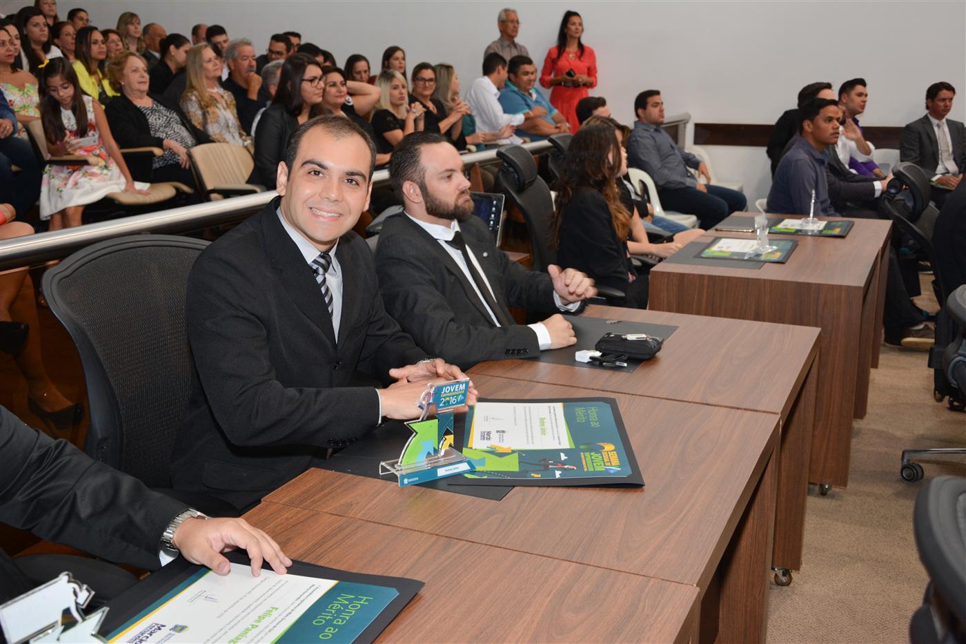 Rodney sorri sentado no plenário da Assembleia Legislativa, com o Troféu Jovem Empreendedor 2016 e um certificado de Honra ao Mérito sobre a mesa